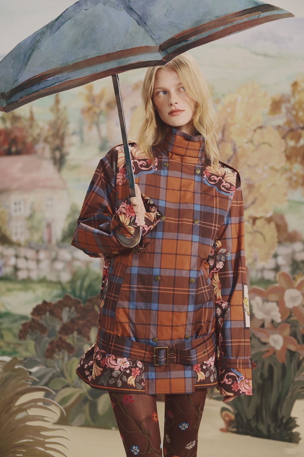 Model holding an umbrella, wearing a belted brown and blue tartan jacket with floral and banana print panels