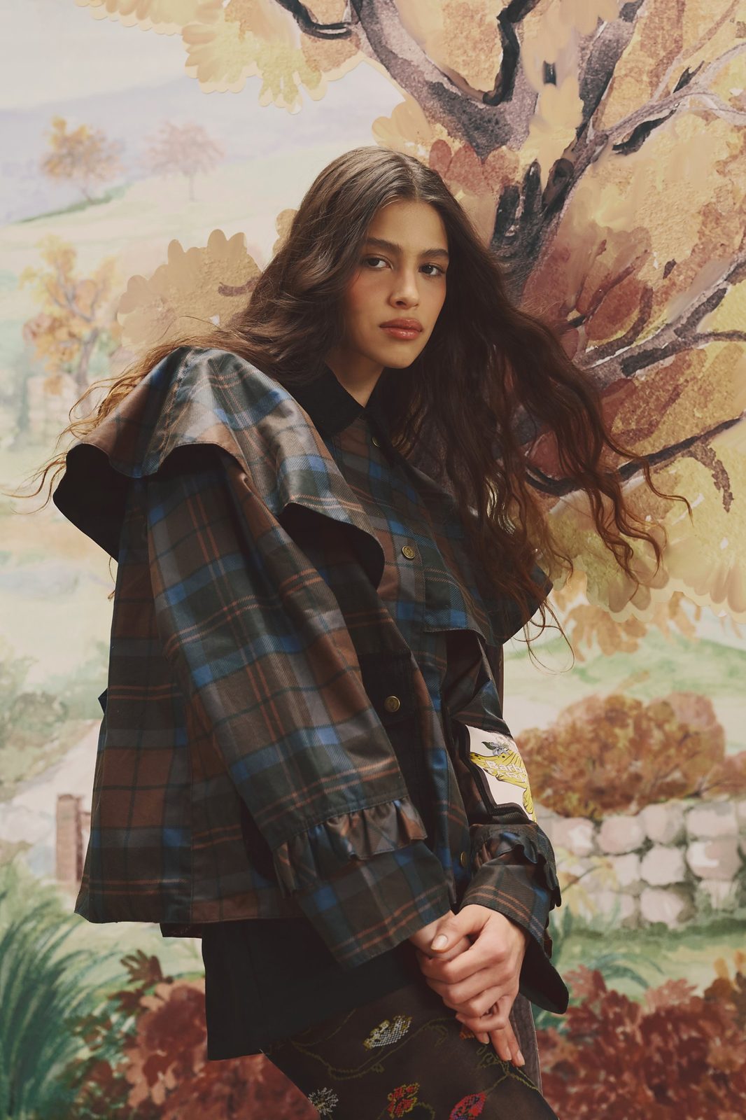 Model in a tartan jacket with a ruffled cape collar and frilled cuffs, in front of a painted autumn landscape