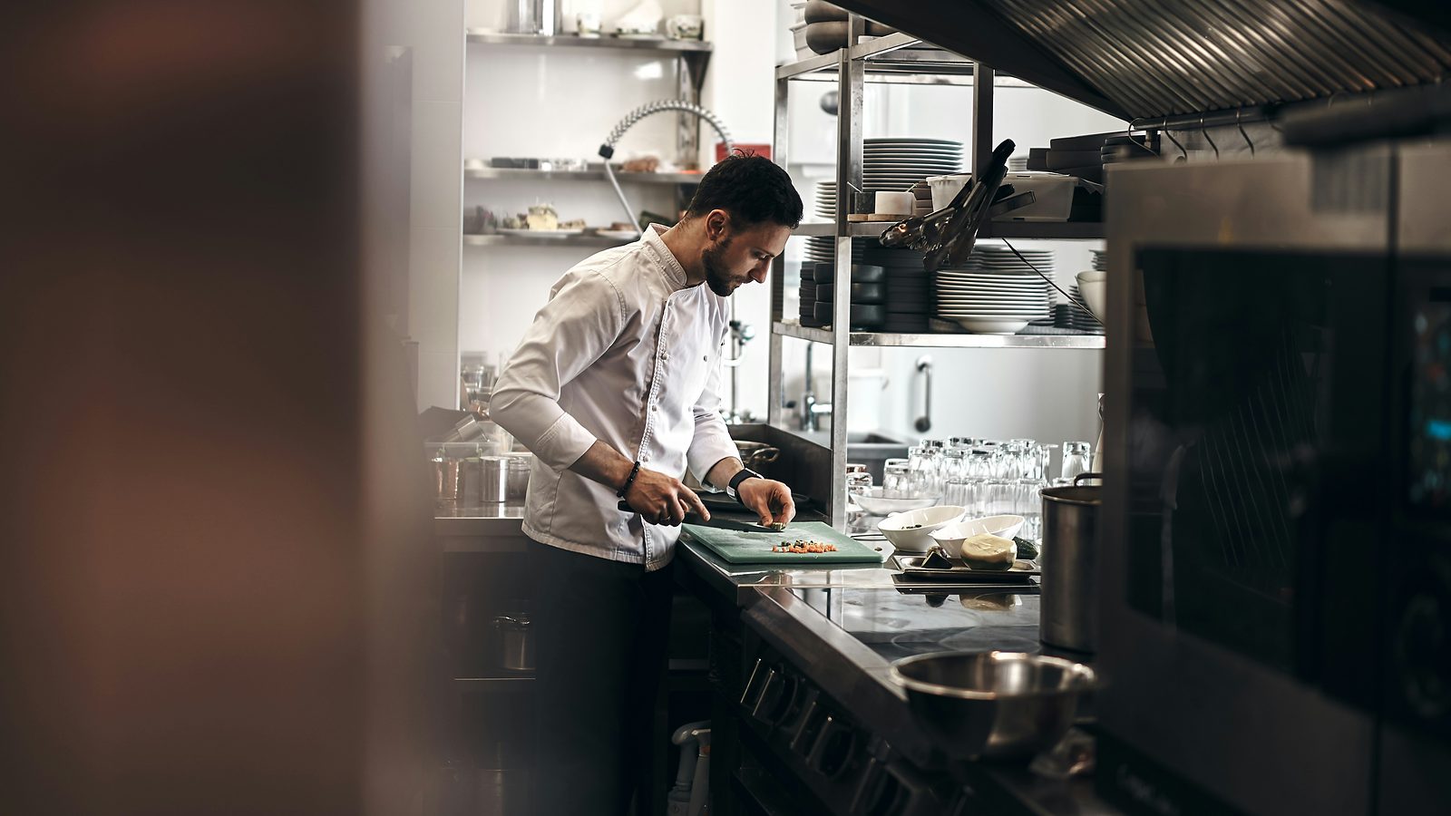 Chef in a white chef's jacket chopping ingredients on a board in a stainless steel restaurant kitchen