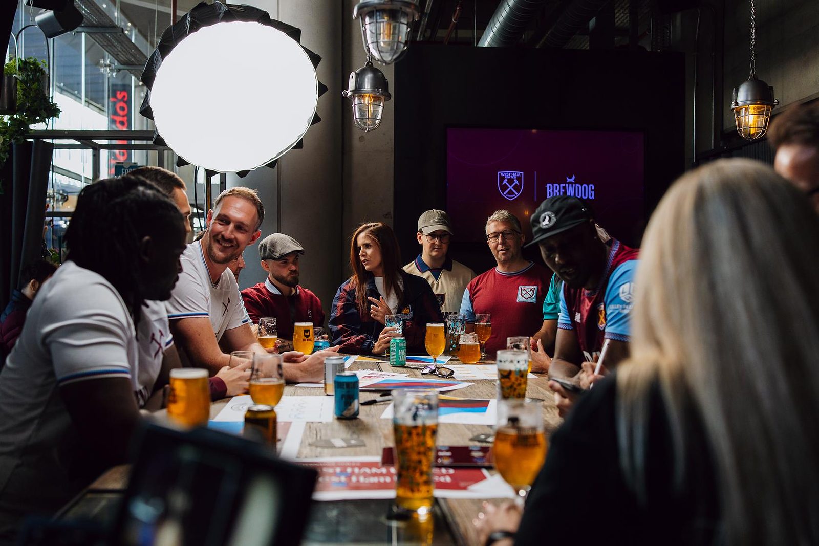West Ham United supporters around a table with beers and design sheets during a Boleyn Brew session, with West Ham United and BrewDog logos on a screen behind